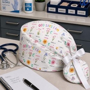 May include: A white scrub cap featuring a colorful floral pattern and the repeating text "GOD SAYS I AM" alongside various Bible verses like "1 THESS 14", "LUKE 7.4", and "GENESIS 1.31". The cap includes a white ribbon tied in a bow at the back.
