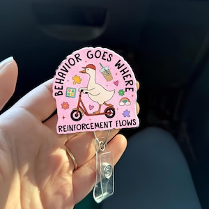 Puede incluir: Un portatarjetas rosa con el texto "BEHAVIOR GOES WHERE REINFORCEMENT FLOWS". La insignia presenta un ganso de dibujos animados montado en un scooter, con café, un arcoíris y otros gráficos.