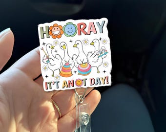 Könnte beinhalten: Ein weißes Badge-Reel mit einem farbenfrohen Design, das Cartoon-Gänse mit Pinseln und Spielzeug zeigt. Der Text "HOORAY IT'S ANOT DAY!" wird mit Blumen- und Smiley-Akzenten angezeigt. Das Reel hat einen transparenten Kunststoffclip.