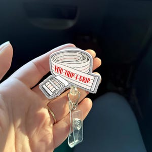 Puede incluir: Un carrete de insignia blanco y rojo con el texto "YOU-TRIP-I-GRIP" en un diseño de cinta métrica blanca. El carrete de insignia tiene un clip de plástico transparente y un cordón retráctil. El carrete de insignia se sostiene en una mano.
