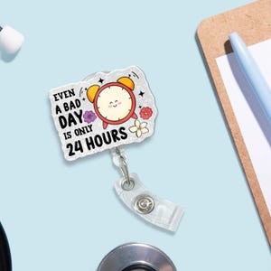 May include: A badge reel featuring a cartoon alarm clock design. The clock is red with yellow bells and a smiling face. The text reads "Even a bad day is only 24 hours." The reel has a clear plastic strap and a metal clip.