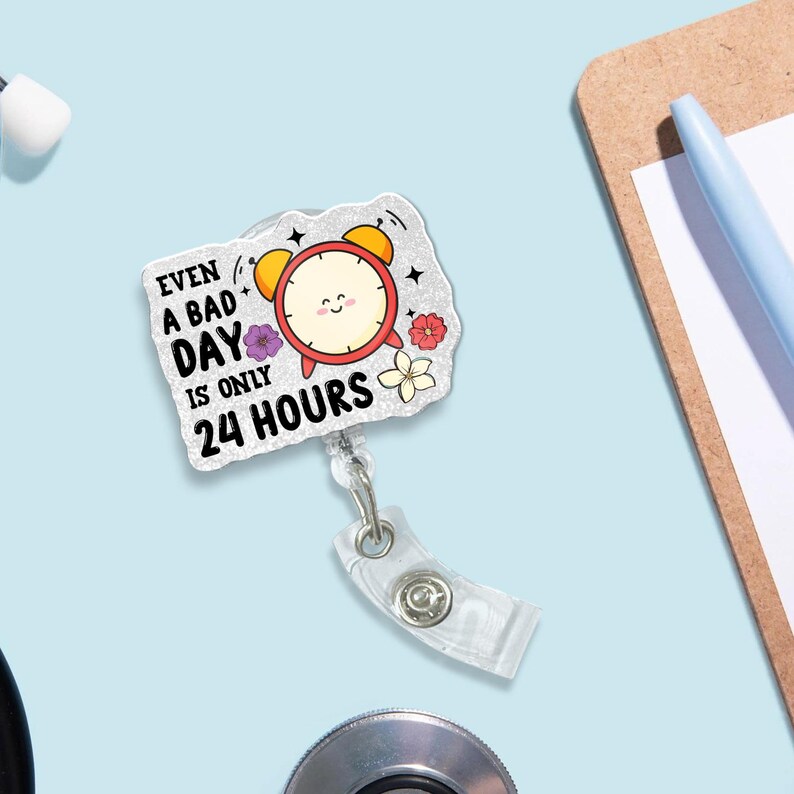 May include: A badge reel featuring a cartoon alarm clock design. The clock is red with yellow bells and a smiling face. The text reads "Even a bad day is only 24 hours." The reel has a clear plastic strap and a metal clip.