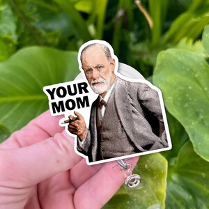 Puede incluir: Un portatarjetas blanco con una pegatina que representa a un hombre con traje sosteniendo un cigarro. El texto "YOUR MOM" est&aacute; impreso en el lado izquierdo de la pegatina. El fondo es un follaje verde borroso.