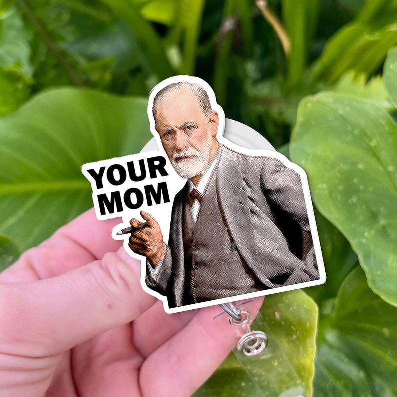 Puede incluir: Un portatarjetas blanco con una pegatina que representa a un hombre con traje sosteniendo un cigarro. El texto "YOUR MOM" est&aacute; impreso en el lado izquierdo de la pegatina. El fondo es un follaje verde borroso.