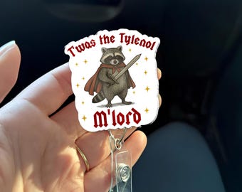 May include: A white badge reel with a cartoon raccoon wearing a cape and holding a sword. The text on the badge reads "Twas the Tylenol" and "m'lord" in red lettering. The reel has a clear plastic retractable cord and a metal clip.