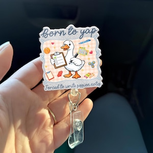 May include: A badge reel with a cartoon goose holding a clipboard and pen. The badge has the text "Born to yap" and "Forced to write session notes". The background is a light pink plaid pattern with various small illustrations.