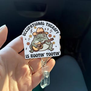 May include: A badge reel with a cartoon frog playing a guitar, wearing a cowboy hat. The badge reel has the text "OCCUPATIONAL THERAPY IS ROOTIN' TOOTIN'". The badge reel is white with a clear plastic clip.
