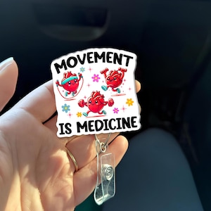 May include: A white badge reel with the words "MOVEMENT IS MEDICINE" and cartoon hearts exercising. The hearts are red with smiling faces and are shown jumping rope, running, and lifting weights. The reel has a clear plastic ID holder.
