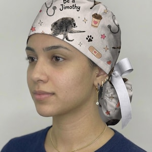 May include: A light-colored scrub cap featuring a repeating pattern of raccoons, stethoscopes, coffee cups, bandages, and stars. The text "Be a Jimothy" is printed on the front. The cap includes a white ribbon tie at the back for adjustment.
