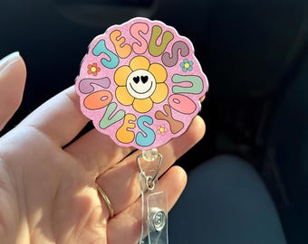 May include: A pink, flower-shaped badge reel with the words "JESUS LOVES YOU" in various colors. The center features a smiling flower with heart-shaped eyes. The reel has a clear plastic clip and a metal retractable cord.
