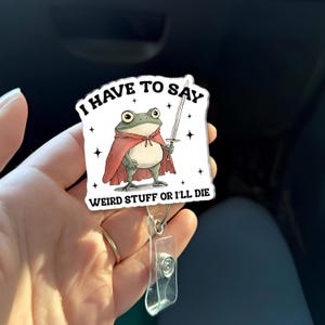 May include: A white badge reel with a cartoon frog wearing a red cape and holding a sword. The text on the badge reads "I HAVE TO SAY WEIRD STUFF OR I'LL DIE". The frog has large eyes and a neutral expression.