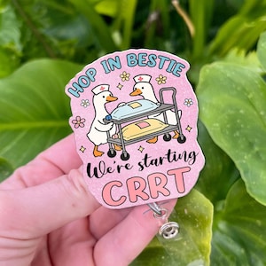 May include: A pink badge reel with the text "HOP IN BESTIE" and "We're starting CRRT". The badge features two cartoon ducks in nurse hats pushing a medical cart. The reel has a clear plastic strap and a metal clip.