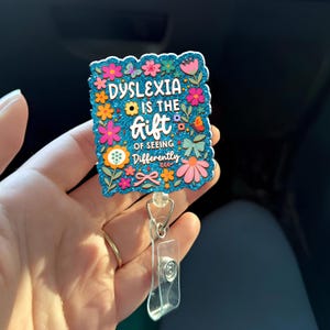 Peut inclure: Un porte-badge avec un fond bleu pailleté et un motif floral. Le texte sur le porte-badge se lit « La dyslexie est le don de voir différemment ». Le porte-badge a un clip en plastique transparent et un cordon rétractable.