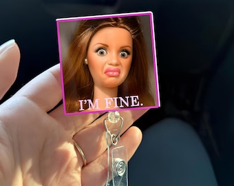 May include: A retractable badge holder with a pink-framed square featuring a doll's face with a distressed expression. The text "I'M FINE." is printed below the image. The holder has a clear plastic clip and a metal clasp.