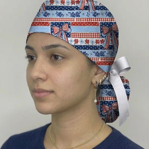 May include: A scrub cap featuring a patriotic pattern of red, white, and blue stripes, polka dots, floral motifs, and bows. The cap is secured at the back with a solid white ribbon tie, creating a gathered look that fits snugly around the head.