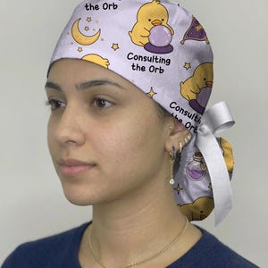 May include: A light purple scrub cap featuring a repeating pattern of yellow chicks looking into purple crystal balls, crescent moons, stars, and potion bottles. The text "Consulting the Orb" is printed in black, and a white ribbon ties at the back.