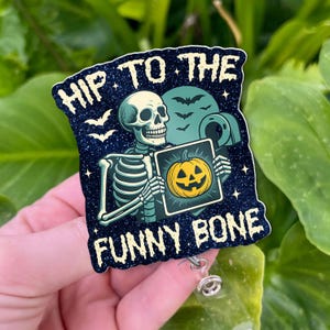 Könnte beinhalten: Ein Abzeichenhalter zum Thema Halloween mit dem Text "HIP TO THE FUNNY BONE". Das Design zeigt ein Skelett, das einen Kürbis hält, Fledermäuse und einen dunkelblauen Hintergrund mit Glitzer.