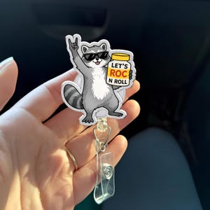 May include: A badge reel featuring a cartoon raccoon wearing sunglasses and giving a rock-and-roll hand gesture. The raccoon is holding a jar with the text "LET'S ROC N ROLL". The reel has a clear plastic clip and a retractable cord.