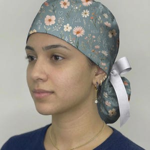 May include: A scrub cap in a muted blue-green color featuring a repeating pattern of pink and white flowers, small green leaves, white hearts, crosses, and the word faith written in a cursive script, finished with a white ribbon tie at the back.
