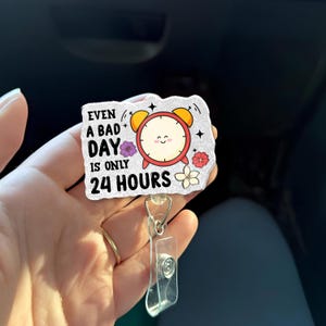 May include: A badge reel featuring the phrase "Even a bad day is only 24 hours." The design includes a cartoon clock, flowers, and stars. The reel is attached to a clear plastic holder with a metal clip.