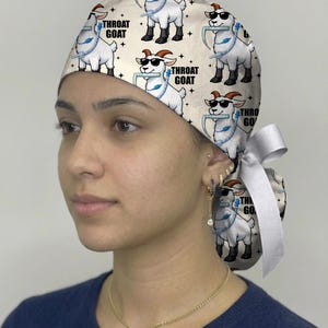 May include: A cream-colored scrub cap featuring a repeating pattern of white goats wearing black sunglasses and holding blue medical laryngoscopes in their mouths. The text "THROAT GOAT" is printed in black next to each goat, and a white ribbon ties at the back.
