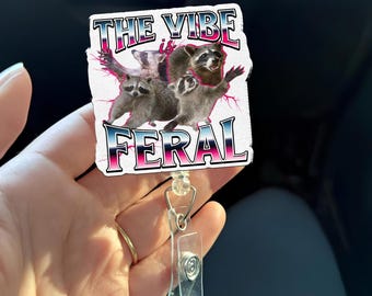 May include: A white retractable badge reel with a graphic of raccoons and the text "THE VIBE is FERAL" in pink and blue. The badge reel has a clear plastic strap and a metal clip.