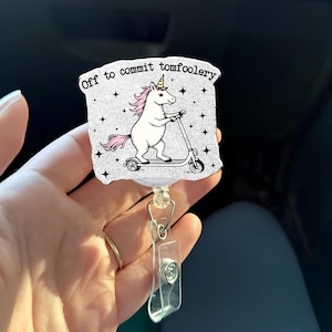 May include: A white badge reel with a cartoon unicorn riding a scooter. The unicorn has a pink mane and tail, and wears a party hat. The text "Off to commit tomfoolery" is printed above the unicorn.