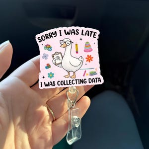 May include: A pink badge reel with the text "SORRY I WAS LATE I WAS COLLECTING DATA". The badge features a cartoon goose wearing glasses and holding a clipboard. Other graphics include a brain, pencil, and flowers.