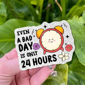 May include: A badge reel with the text "EVEN A BAD DAY IS ONLY 24 HOURS". The badge features a smiling cartoon alarm clock, purple, red, and white flowers, and a glittery background. The reel has a clear plastic retractable cord.