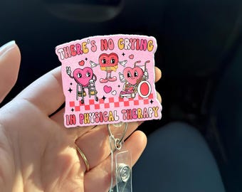 Puede incluir: Un portatarjetas rosa con el texto "THERE'S NO CRYING IN PHYSICAL THERAPY". La insignia presenta personajes de corazón de dibujos animados con varios equipos de fisioterapia. El carrete tiene un clip de plástico transparente y un anillo de metal.