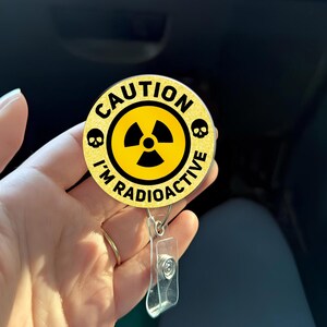 May include: A round, yellow and black badge with the words "CAUTION I'M RADIOACTIVE" in a circular design. The badge features a radiation symbol in the center and two skull icons. It has a clear plastic retractable badge holder.