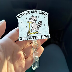 Op de afbeelding: Een lichtblauwe badge reel met de tekst "BEHAVIOR GOES WHERE REINFORCEMENT FLOWS". De badge reel toont een cartoon wasbeer op een gele scooter. De badge reel heeft een doorzichtige plastic clip en een metalen ring.