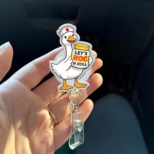 May include: A white goose wearing a nurse's cap with a red cross, holding a jar with the text "LET'S ROC N ROLL". The badge reel has a clear plastic strap and a metal clip.