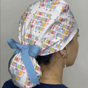 May include: A white scrub cap with a colorful text design that reads "I don't get distracted, I go on side quests." The cap is tied with a light blue bow. The design includes flowers and stars.