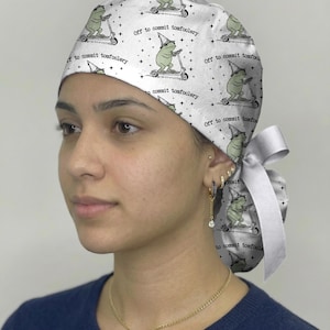 May include: A white scrub cap featuring a repeating pattern of green frogs wearing pointed hats and riding scooters, accompanied by the text "Off to commit tomfoolery" and small black stars. The cap includes a white ribbon tie at the back.