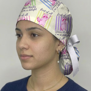 May include: A scrub cap featuring a repeating pattern of pink raccoons and vertical stripes in yellow, blue, and purple. The text "first of all" and "Jesus thinks I'm a delight" is printed across the fabric. A white ribbon is tied at the back.