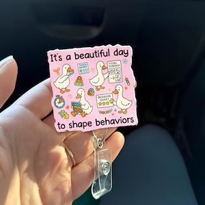 Puede incluir: Una insignia rosa con el texto "It's a beautiful day to shape behaviors" e ilustraciones de patos con gráficos de comportamiento y horarios visuales. La insignia tiene un clip de plástico transparente.