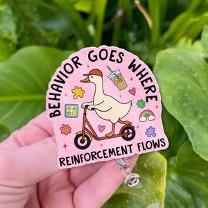 K&ouml;nnte beinhalten: Ein rosa Ausweisrolle mit dem Text "BEHAVIOR GOES WHERE REINFORCEMENT FLOWS". Das Design zeigt eine Gans auf einem Roller, ein Kaffeegetr&auml;nk und andere farbenfrohe Grafiken. Die Rolle hat einen transparenten Kunststoffclip.
