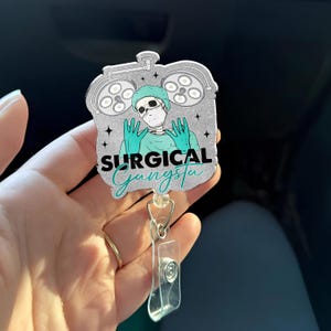 Op de afbeelding: Een zilveren badge reel met een cartoon skelet met een chirurgische muts, masker en handschoenen, met chirurgische lampen op de achtergrond. De tekst "SURGICAL Gangsta" is in teal.