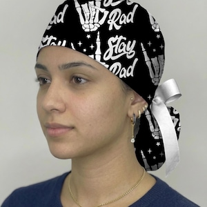 May include: A black scrub cap featuring a white repeating pattern of skeleton hands making a rock-and-roll gesture, small stars, and the text "Stay Rad" in a cursive font. The cap includes a white ribbon tie at the back for an adjustable fit.