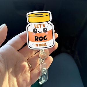 Może przedstawiać: Biało-pomarańczowy uchwyt na identyfikator z napisem "LET'S ROC N ROLL". Uchwyt na identyfikator ma kreskówkowy wzór butelki z lekarstwem z żółtą nakrętką i przezroczystym plastikowym klipsem.