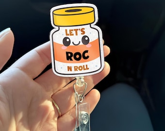 May include: A white and orange badge reel with the text "LET'S ROC N ROLL" in black. The badge reel has a cartoon design of a medicine bottle with a yellow cap and a clear plastic clip.