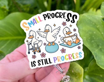 Könnte beinhalten: Ein weißes Ausweis-Jojo mit einem Cartoon-Enten-Design. Das Design zeigt drei Enten in verschiedenen Posen mit dem Text "SMALL PROGRESS IS STILL PROGRESS" in einem Regenbogenfarbschema. Das Ausweis-Jojo hat einen transparenten Kunststoffhalter und einen Metallclip.