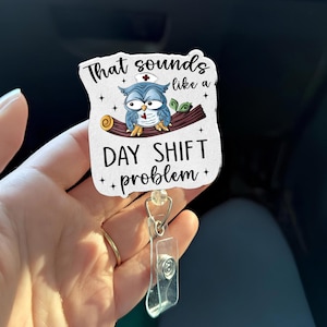 Könnte beinhalten: Ein weißes Ausweis-Jojo mit dem Text "That sounds like a DAY SHIFT problem" und einer Cartoon-Eule mit Krankenschwestermütze. Die Eule sitzt auf einem braunen Ast. Das Jojo hat einen transparenten Kunststoffhalter und einen Metallclip.