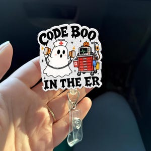 May include: A glittery badge reel featuring a white ghost dressed as a nurse holding defibrillator paddles next to a red medical cart with a pumpkin on top. The text reads CODE BOO IN THE ER in black, dripping, stylized lettering.