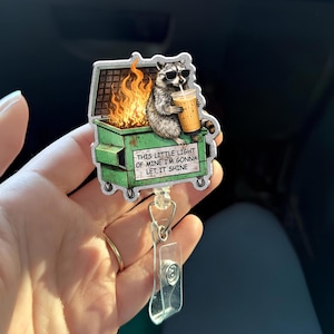 May include: A badge reel featuring a raccoon wearing sunglasses, holding a drink, and sitting on a dumpster with flames. The badge reel has the text "THIS LITTLE LIGHT OF MINE I'M GONNA LET IT SHINE". The reel has a clear plastic clip.