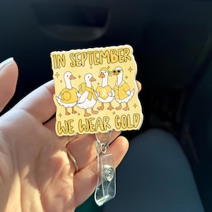 May include: A badge reel with a yellow and white design featuring four cartoon geese wearing yellow accessories. The text "IN SEPTEMBER WE WEAR GOLD" is printed on the badge. The reel has a clear plastic clip and a metal retractable cord.