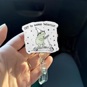 May include: A white badge with a cartoon frog riding a scooter, wearing a wizard hat. The badge has the text "Off to assess behavior" and is attached to a clear plastic retractable holder.