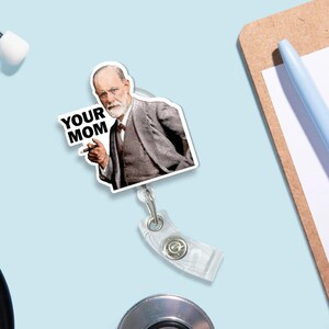 Puede incluir: Un portatarjetas blanco con una imagen impresa de un hombre con traje se&ntilde;alando, con el texto "YOUR MOM". El portatarjetas tiene un cord&oacute;n retr&aacute;ctil de pl&aacute;stico transparente y un clip met&aacute;lico.