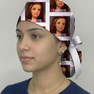 Peut inclure: Une charlotte chirurgicale blanche avec un motif r&eacute;p&eacute;titif repr&eacute;sentant une poup&eacute;e aux cheveux bruns &agrave; l'expression contrari&eacute;e, encadr&eacute;e dans des carr&eacute;s magenta avec le texte I'M FINE. imprim&eacute; en blanc sous chaque image. Ruban blanc.